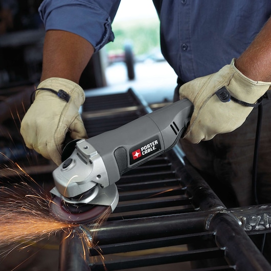 A close-up view of a person wearing gloves using a PORTER-CABLE angle grinder to cut or grind a metal bar, with sparks visible from the grinding process.