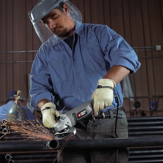 A person wearing protective gloves and a face shield is using a PORTER-CABLE angle grinder (model PC750AG) to cut a metal pipe, creating sparks, inside an industrial workshop.