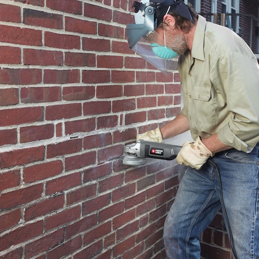 A person wearing protective gear, including a face shield, mask, and gloves, uses a PORTER-CABLE angle grinder to work on the mortar joints of a brick wall.