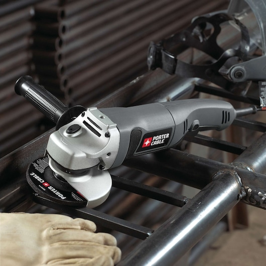 A PORTER-CABLE angle grinder, model PC750AG, is shown on a metal structure with a gloved hand nearby, likely during a metalworking operation. The tool features a black and silver design and is plugged in for use.
