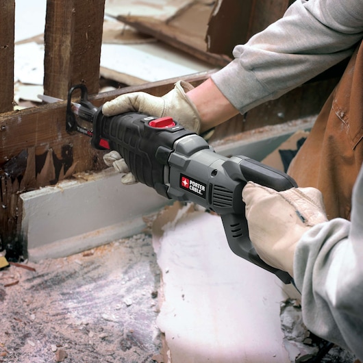 A person wearing gloves is using a PORTER-CABLE reciprocating saw to cut through wooden studs during a demolition or construction project. The area appears to be covered with debris and dust.