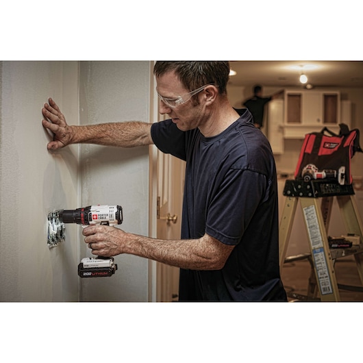 A person using a PORTER-CABLE cordless drill to install or adjust an electrical outlet in a wall. The drill shows the brand and 20V lithium battery. In the background, there is a ladder and a tool bag on a table in a home improvement setting.