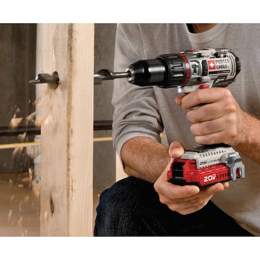 A person is using a PORTER-CABLE 20V lithium cordless drill to bore a hole into a wooden beam, with wood shavings flying out from the hole.