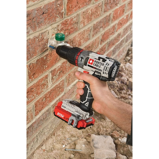 A person uses a PORTER-CABLE 20V lithium cordless drill to drill into a brick wall next to a green metal pipe fitting. The drill is held in one hand and is being used outdoors against a red brick surface.