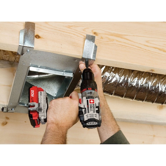 A PORTER-CABLE cordless drill (model PCC620LB) is being used to fasten a metal duct to a wooden beam, with a person's hands holding the tool and securing the metal fitting. The drill features a 20V lithium-ion battery.