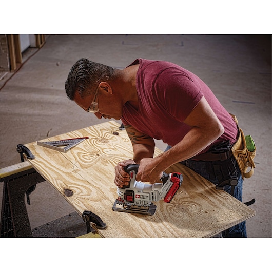A person using a PORTER-CABLE cordless jigsaw (model PCC650B) to cut a piece of plywood, with the wood clamped to a workbench. The person is wearing a tool belt and a maroon shirt. A square measuring tool is also visible on the plywood.
