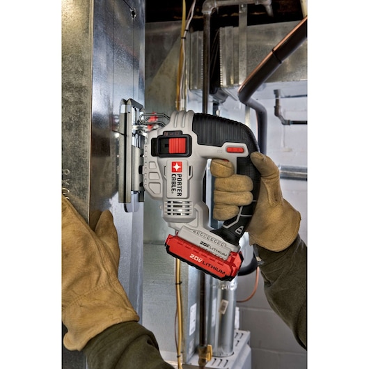 A person wearing work gloves is using a PORTER-CABLE 20V Lithium cordless jigsaw to cut through a metal sheet in an industrial or workshop setting.