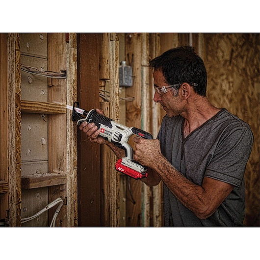 A person using a PORTER-CABLE PCC670B cordless reciprocating saw with a 20V lithium battery to cut through wooden framing at a construction site. The person's face is blurred for privacy.