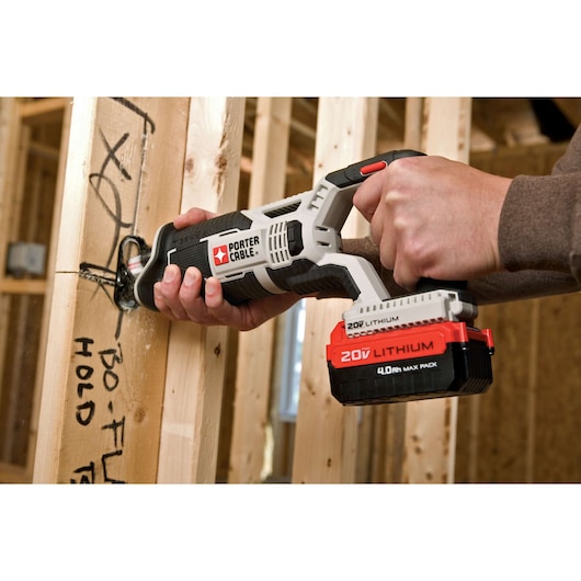 A person using a PORTER-CABLE 20V lithium cordless reciprocating saw to cut through a wooden stud in a construction setting. The saw is black, white, and red, with a visible PORTER-CABLE logo and 20V lithium battery attached. The wooden stud has black marker writing on it.