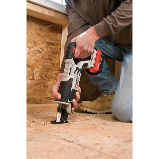 A person uses a PORTER-CABLE cordless reciprocating saw, model PCC670B, to cut wood flooring inside a construction area. The saw has a red and black 20V lithium-ion battery attached.