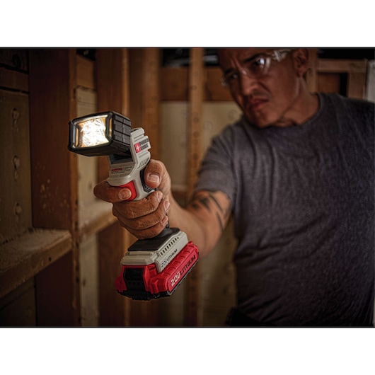 A person holding a PORTER-CABLE PCC700B cordless LED work light with a 20V lithium-ion battery, used for illumination in a construction or workshop setting. The person's face is blurred.