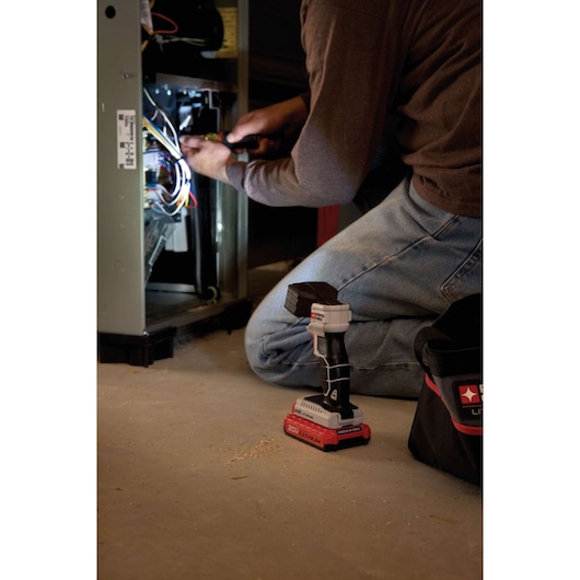 A PORTER-CABLE cordless LED flashlight (model PCC700B) with a 20V lithium battery is shown on the floor near a tool bag, while a person kneels and works on wiring inside an open electrical panel.