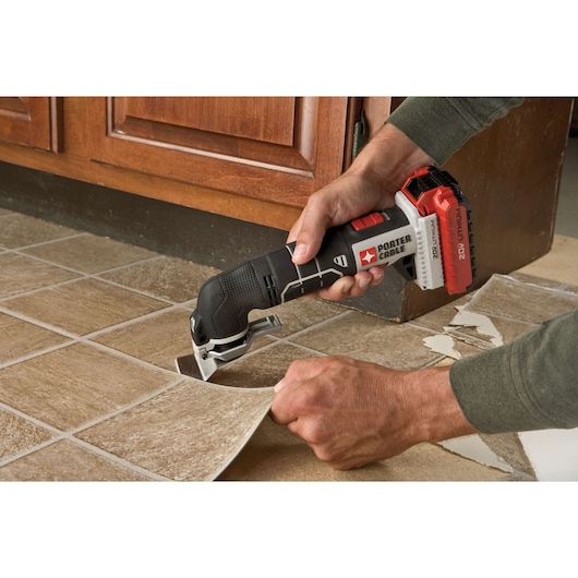 A person uses a PORTER-CABLE cordless oscillating multi-tool (model PCC710B) to remove tile flooring in a kitchen setting. The tool is cutting through adhesive under a tile piece, helping lift and separate it from the floor.