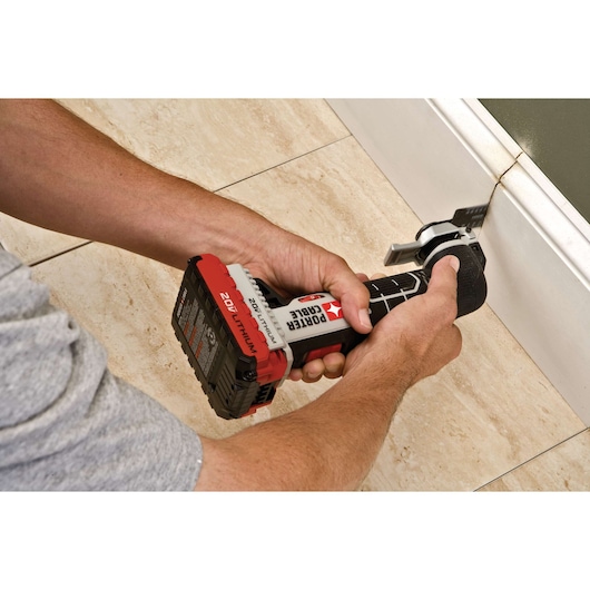 A person using a PORTER-CABLE 20V cordless oscillating multi-tool to cut along the edge of a tiled floor near a baseboard.
