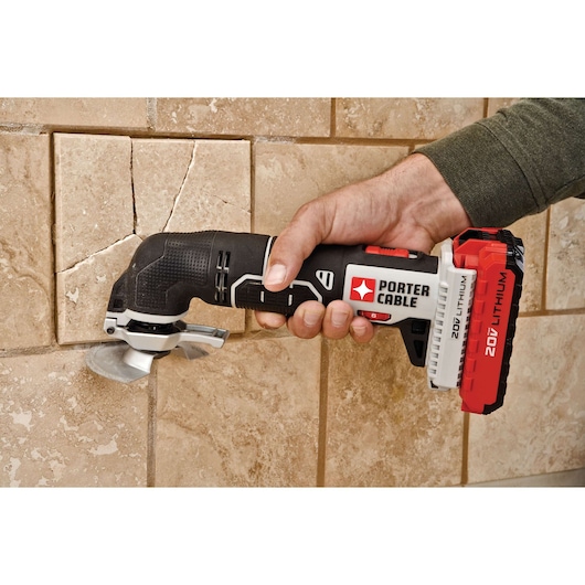 A hand holding a PORTER-CABLE PCC710B cordless oscillating multi-tool with a 20V lithium battery, being used to cut or remove a cracked tile from a wall.
