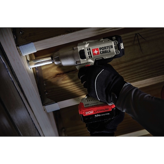 A person wearing black gloves is using a PORTER-CABLE 20V lithium cordless impact wrench to tighten a bolt under a wooden structure. The tool is branded with the PORTER-CABLE logo and features an LED light.