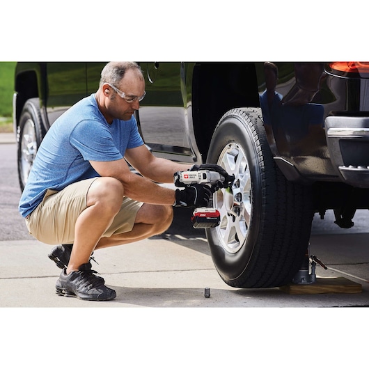 A person wearing gloves uses a PORTER-CABLE cordless impact wrench (model PCC740LA) to remove or tighten the lug nuts on a car wheel. The vehicle is raised with a jack and the person is kneeling on the ground outdoors.