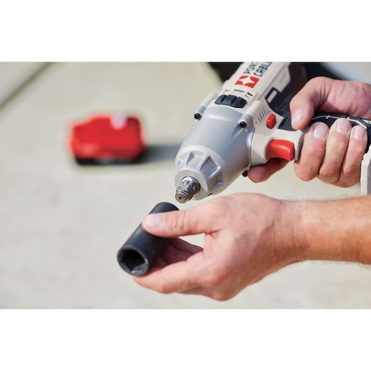 A person holds a PORTER-CABLE cordless impact wrench and a socket head, preparing to attach the accessory. The background includes a red battery pack placed on a concrete surface.