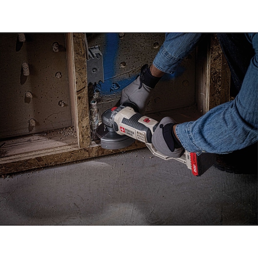 A person wearing gloves and a denim shirt uses a PORTER-CABLE cordless power tool, likely an angle grinder, to cut or grind materials at a construction site near a wooden frame and wall.