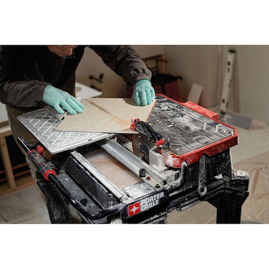 A person wearing gloves is using a PORTER-CABLE tile saw to cut a ceramic tile in a workshop setting. The saw features a red and black design and the workspace is covered with dust and construction materials.