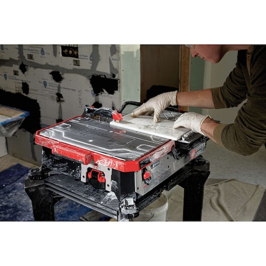 A person wearing gloves operates a PORTER-CABLE tile saw, cutting a piece of tile on the worktable. The saw and workspace have visible dust from tile cutting, and the background shows a construction area.
