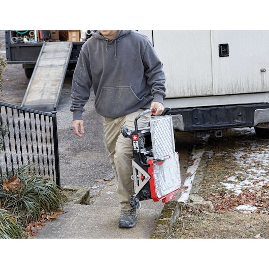 A person wearing a gray hoodie and khaki pants carries a folded PORTER-CABLE tile saw (SKU: PCC780LA) on a sidewalk, with a work truck and ramp in the background. The tile saw is portable and compact for easy transportation.