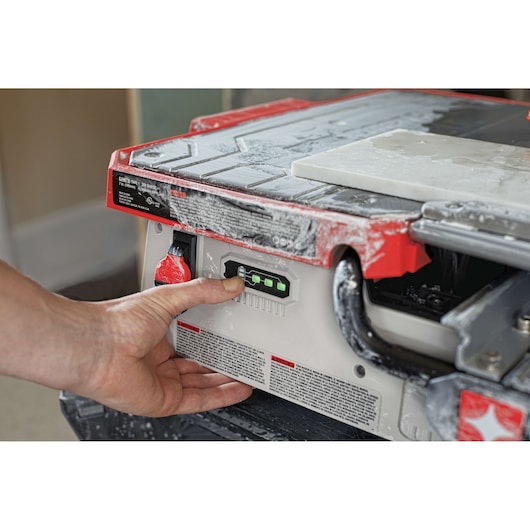Close-up of a person's hand operating the control panel on a PORTER-CABLE tile cutting machine in a photo studio. The tile cutting machine, covered in dust, is actively being used with a tile positioned on the cutting surface.