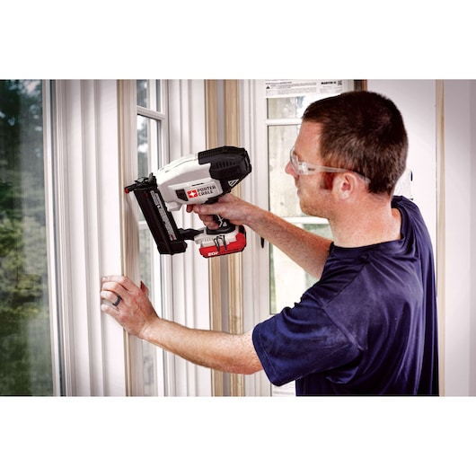A person using a PORTER-CABLE cordless nailer (model PCC790LA) on window trim during a construction or installation project. The individual is wearing safety glasses and holding the tool against the trim.
