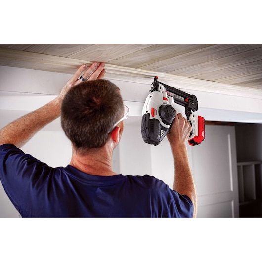 A person is using a PORTER-CABLE cordless nail gun to attach wooden trim to a ceiling. The individual is holding the nail gun with one hand and positioning the wood with the other hand.
