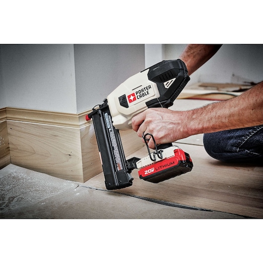 A person is using a PORTER-CABLE 20V Lithium cordless nail gun to attach trim to the base of a wall.