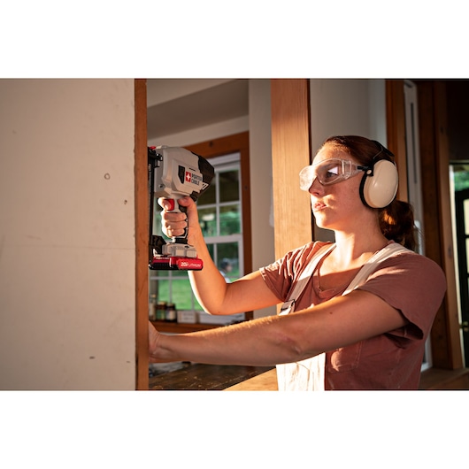 A person wearing ear protection uses a PORTER-CABLE 20V lithium cordless nail gun to install drywall inside a home. The person's face is blurred for privacy.