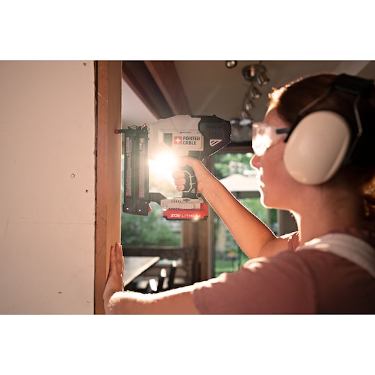 A person wearing safety goggles and ear protection is using a PORTER-CABLE 20V lithium cordless nailer to fasten wood trim to a wall in a well-lit indoor space.