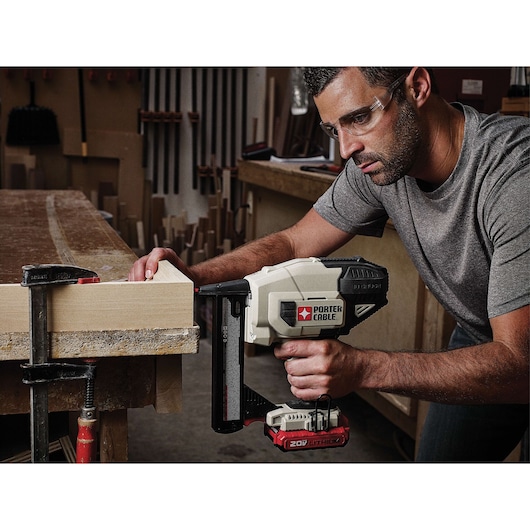 A person is using a PORTER-CABLE 20V lithium cordless nail gun to secure a piece of wood clamped to a workbench in a woodworking shop. The person's face is obscured, and the environment shows various woodworking tools and materials.