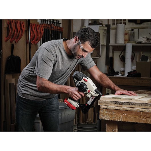 A person using a PORTER-CABLE PCC791LA nail gun on a piece of wood in a workshop. The individual's face is blurred, and various tools and materials are visible in the background.