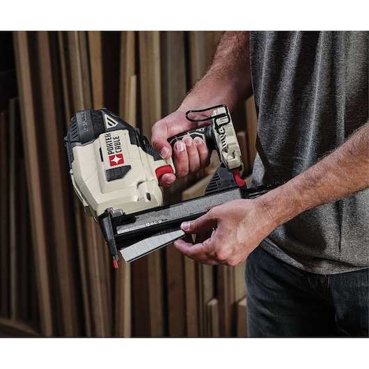A person is holding a PORTER-CABLE cordless nail gun, model PCC791LA, in a woodworking environment with wooden planks in the background.