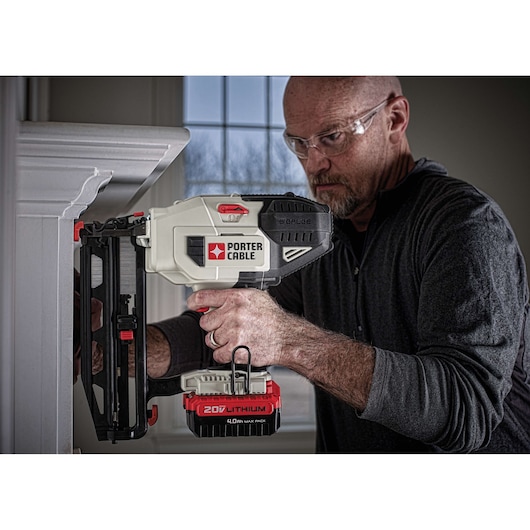 A person is using a PORTER-CABLE 20V lithium cordless nail gun to attach trim to a wall. The person's face is obscured for privacy. The tool and its battery are clearly visible.