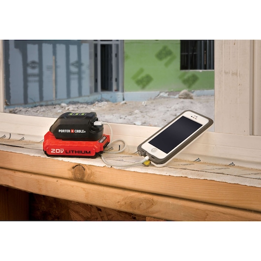A PORTER-CABLE 20V lithium battery with a charging adapter is connected to a smartphone, placed on a windowsill at a construction site.
