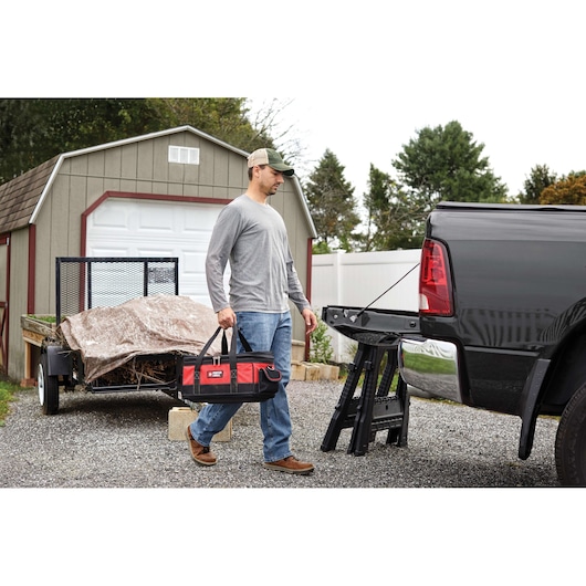 A person carrying a PORTER-CABLE branded red and black tool bag walks near a parked pickup truck with its tailgate down and a folded loading ramp, in an outdoor setting with a shed and trailer in the background.