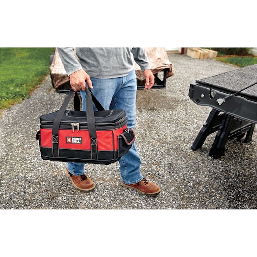 A person holding a PORTER-CABLE branded red and black tool bag with a zippered top, standing outdoors on a gravel surface next to a vehicle and some equipment.