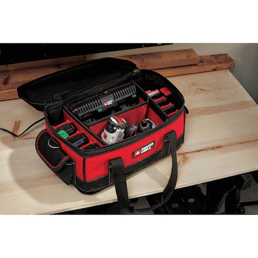A red and black PORTER-CABLE branded tool bag is open on a wooden work surface, revealing compartments filled with various tools and accessories including a battery charger, batteries, and a drill.