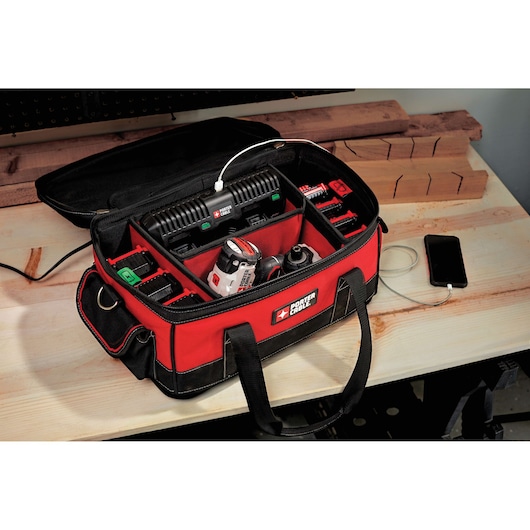 A PORTER-CABLE tool bag containing a battery charger, cordless drill, batteries, and other accessories is placed on a wooden workbench. The bag is red and black, and a smartphone is connected to the charger via a USB cable.
