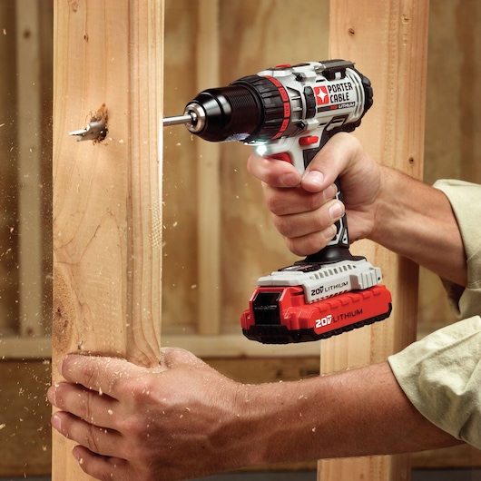 A person uses a PORTER-CABLE 20V Lithium cordless power drill to drill a hole into a wooden beam, with wood shavings flying around.