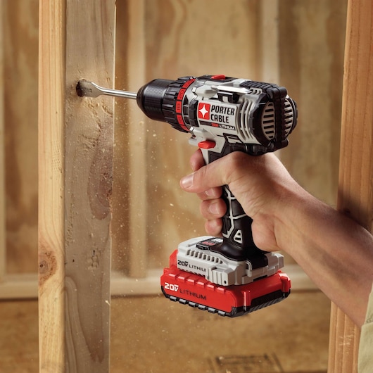 A PORTER-CABLE 20V lithium cordless drill is being used to drill a hole in a wooden beam. Wood dust is visible as the drill bit works through the material. The drill is held by a person indoors, likely at a construction or renovation site.