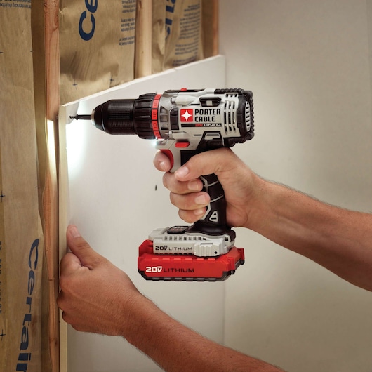 A person is using a PORTER-CABLE 20V lithium cordless drill to drive a screw into drywall during construction. The drill has a built-in light illuminating the work area.
