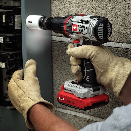 A person wearing protective gloves uses a PORTER-CABLE 20V lithium cordless drill to create a hole in a metal electrical panel mounted on a concrete wall.