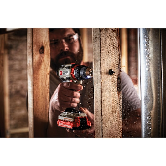 A person using a PORTER-CABLE cordless drill to drill a hole into a wooden beam. Wood shavings are visible as the drill is in action. The focus is on the tool and its application in a construction or woodworking environment.