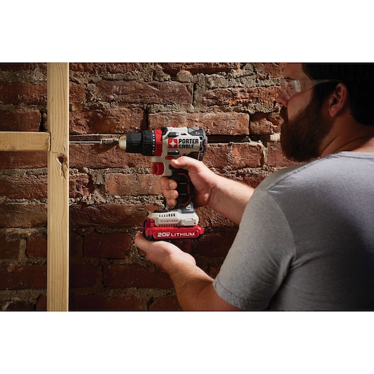 A person using a PORTER-CABLE 20V Lithium cordless drill to drill into a wooden beam, with a brick wall in the background.