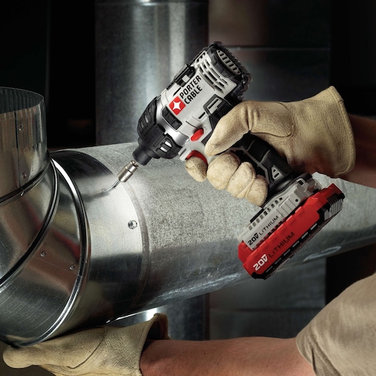 A person wearing work gloves uses a PORTER-CABLE 20V lithium cordless power drill to fasten a screw into a metal duct.