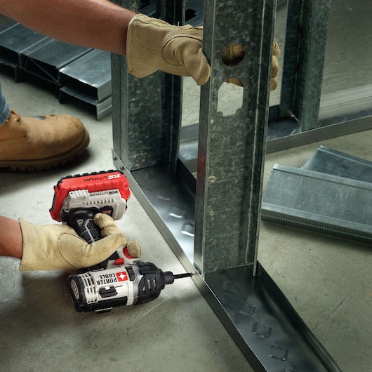 A person wearing gloves and work boots uses a PORTER-CABLE cordless drill to secure a metal stud frame to the floor, with various metal sections visible nearby.
