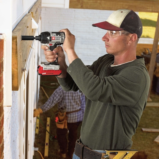 A person using a PORTER-CABLE 20V Lithium cordless power drill to fasten screws into wood on a construction site, with another worker and tools visible in the background. The person's face is blurred for privacy.